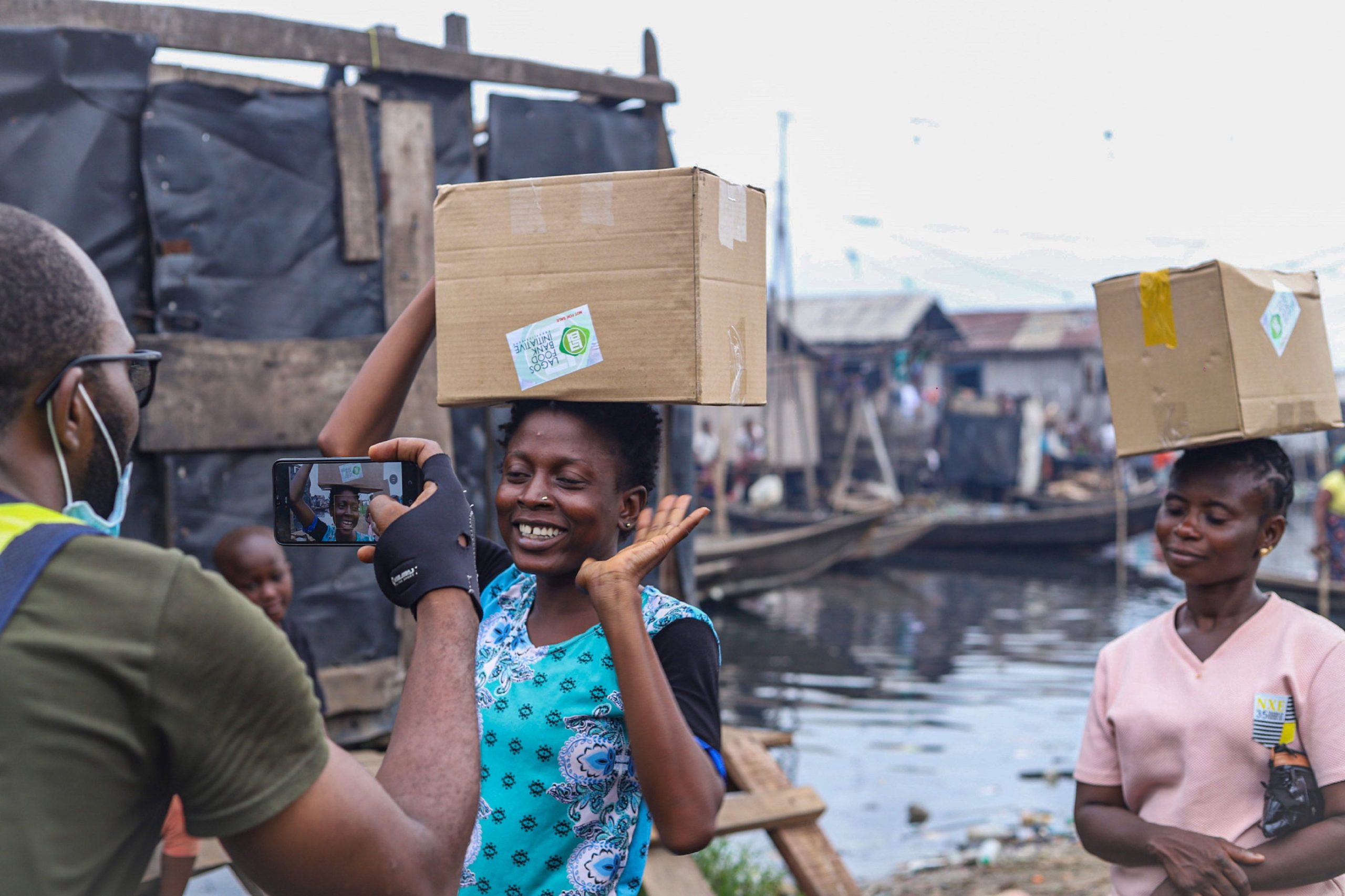Two women carrying boxes on their heads pose for a photo outdoors by waterfront.
